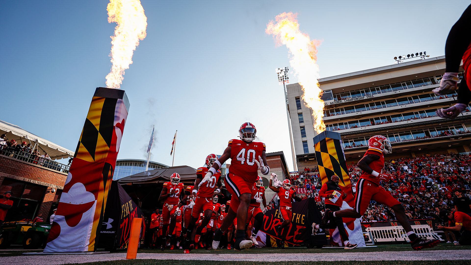 The Maryland Football team runs onto the field at SECU Stadium through two Maryland flag towers with a fiery display.